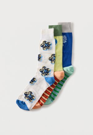 Trois chaussettes montantes colorées empilées : blanche avec des imprimés floraux bleus et un bout bleu, jaune avec un revers vert et un pied rayé orange, bleue unie avec un revers gris.