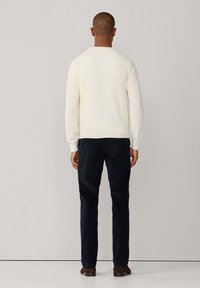 Mann, der einer Wand gegenübersteht, trägt einen weißen Pullover mit langen Ärmeln, dunkle Hosen und braune Schuhe vor einem neutralen Hintergrund.