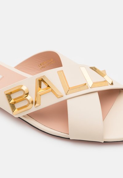 Beige leather sandals feature a crisscross design with a prominent gold "BALLY" logo on the strap. The interior is a soft pink.