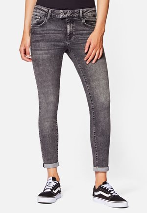 Personne portant un jean skinny noir délavé avec des revers roulés et des baskets Vans noir et blanc, debout devant un fond uni.