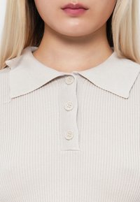Beige ribbet strikket polo skjorte med krage, som har fire matchende knapper langs pløyten og en jevn tekstur.