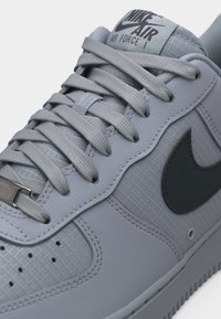 Lähikuva harmaasta Nike Air Force 1 -tossusta, jossa on kuvioitu päällinen, tasaiset nauhat, metallinen nauhanmerkki ja musta Nike Swoosh -logo.