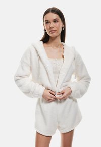 Sudadera blanca y esponjosa con cremallera frontal, cordones ajustables, mangas largas y pantalones cortos a juego. Textura suave y apariencia acogedora.