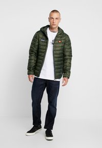 Jachetă pufoasă verde măsliniu cu glugă, textură matlasată, fermoar frontal și detalii cu logo. Purtați-o peste un tricou alb și jeans albaștri.