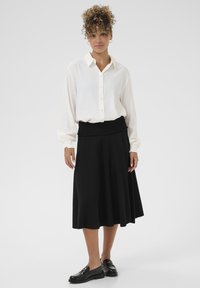 Bijela košulja s dugim rukavima, suknja s crnim pletenim pojasom i sjajne crne mokasinke, stvarajući klasičan i elegantan izgled.