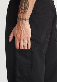 Pantalones negros con un bolsillo lateral y costuras visibles. Mano descansando sobre el bolsillo, con un anillo plateado y un tatuaje en el antebrazo.