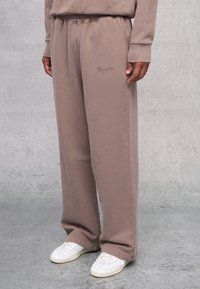 Sudadera de felpa marrón con un corte relajado, que presenta una cinturilla elástica y un logotipo bordado sutil en el muslo. Combinada con zapatillas blancas.