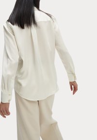 Blusa de seda crema con una textura suave, cuello clásico y puños abotonados, que presenta un bajo redondeado y un corte aéreo.