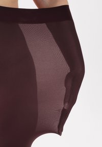 Collant bordeaux trasparent con una consistenza liscia, vita alta e un design aderente, messo in risalto da una mano che afferra il materiale.