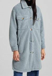 Fuzzy, light blue coat made of textured fabric, featuring a collar, two chest pockets, buttoned front, and long sleeves.
