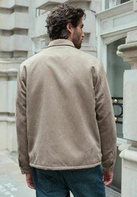 Jachetă bej din suede cu o textură netedă, având un design minimalist, croială ajustată și detalii de cusătură subtile. Vedere din spate prezentată.