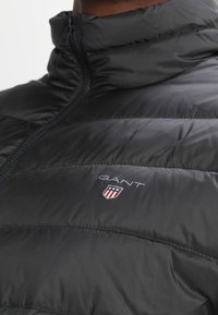 Musta puhvijaakku korkealla kauluksella, jossa on vetoketju edessä ja GANT-logo kilven kanssa. Sileä pinta ja tikattu muotoilu.