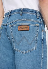 Světle modré džínové kalhoty s koženou nášivkou značky Wrangler na zádech, s výrazným křivým prošitím kapes a úzkým střihem.