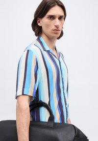 Νεαρός άνδρας με καστανά μαλλιά μέχρι τους ώμους, που φοράει μπλε ριγέ κοντομάνικο πουκάμισο, κρατώντας μια μεγάλη μαύρη δερμάτινη τσάντα ταξιδιού.