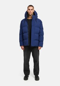 Námornícky modrý páperový kabát s zipsom a patentkami, ktorý má kapucňu a elastické šnúrky, nosený cez čierne tričko a čierne nohavice.