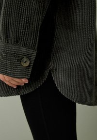 Donkergroene corduroy overhemd met verticale ribbels, knoopmanchetten en een ronde zoom. Draagt over zwarte leggings.
