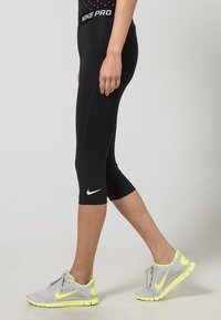 Mustat Nike Pro capri leggings, joissa on valkoinen swoosh-logo, yhdistettynä vaaleanharmaisiin urheilukenkiin, joissa on neonkorostuksia ja karhea pinta.