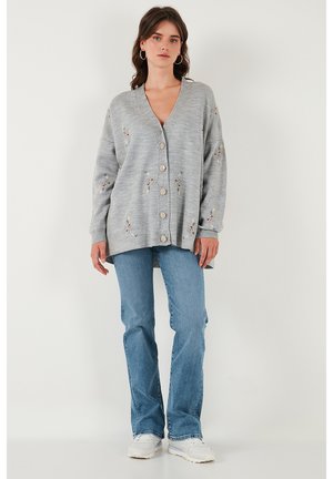 Cardigan grigio chiaro con scollo a V, vestibilità oversize, caratterizzato da ricami floreali, sei bottoni sul davanti e polsini a coste. Indossato con jeans a zampa.