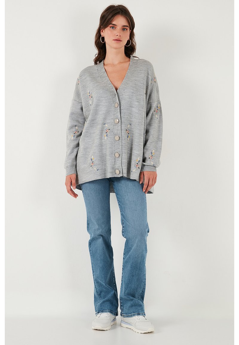 Cardigan gris claro con escote en V, de corte oversized, con bordados florales, seis botones en la parte delantera y puños de canalé. Lleva puesto con jeans acampanados.