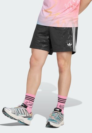 Ben iført sorte Adidas-shorts med hvide striber, lyserøde sokker med teksten "LOVE" og multifarvede sneakers, der står på en lys gulv.