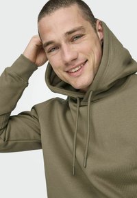 Sudadera color verde oliva con capucha ajustable, puños acanalados y textura de tela suave. El modelo sonríe y toca la capucha con una mano.