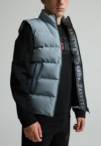 Veste matelassée bleu clair avec un col montant, fermeture éclair noire et détails de logo sur le devant, portée sur un sweatshirt noir et un pantalon assorti.