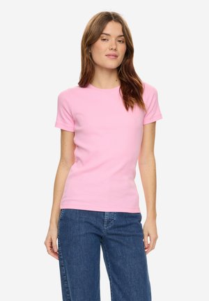 Vrouw draagt een effen lichtroze T-shirt met korte mouwen en een blauwe spijkerbroek, staand tegen een witte achtergrond.