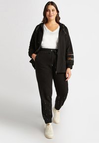 Schwarzer Trainingsanzug aus weichem Material, bestehend aus einer Reisverschluss-Hoodie mit Nietenakzenten und locker sitzenden Jogginghosen mit elastischen Bündchen. Darunter ein weißes T-Shirt.