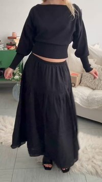 Γυναίκα με μαύρο crop top μακρύ μανίκι και μαύρη φαρδιά φούστα στέκεται σε ένα λευκό σαλόνι με πράσινη ντουλάπα και βιβλία.