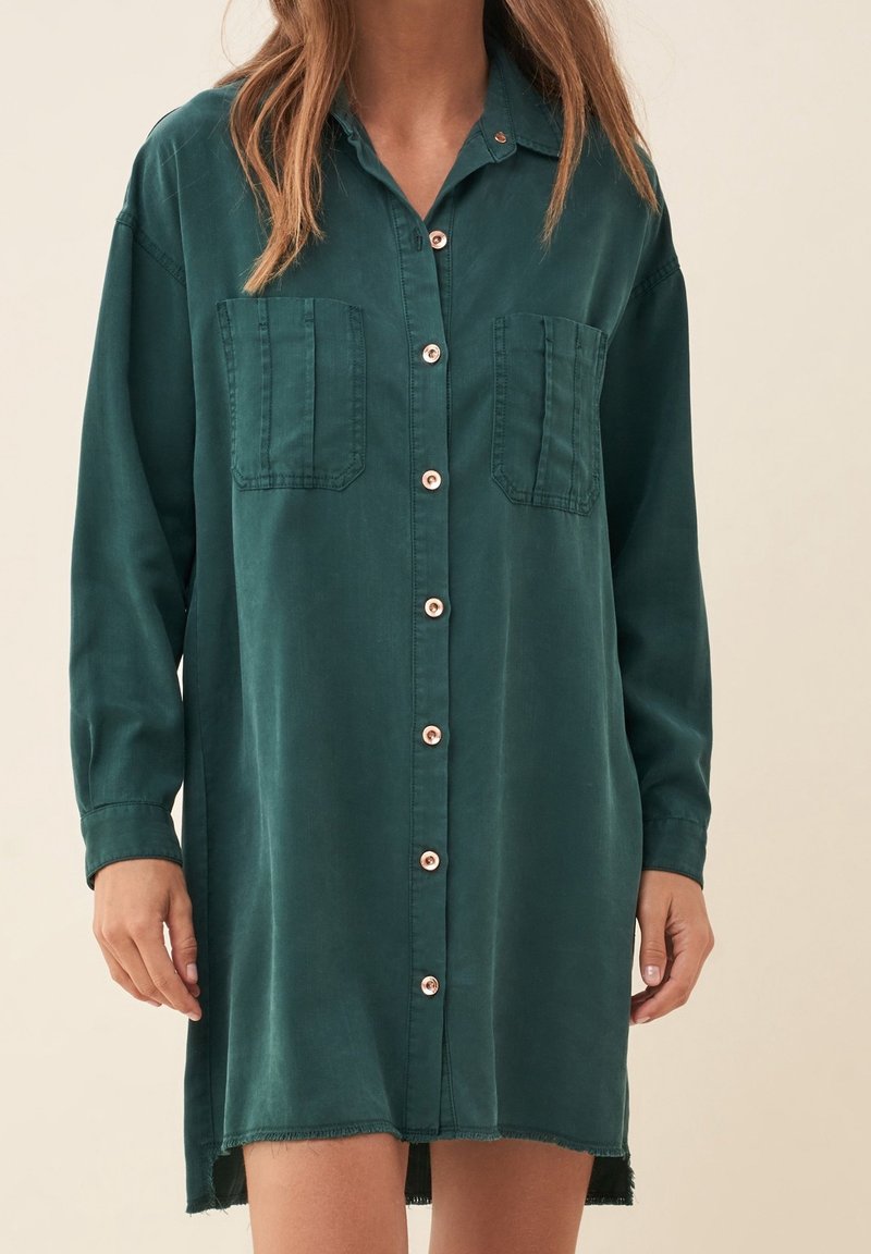 Vestito camicia verde in tessuto morbido, con due tasche sul petto, colletto e orlo sfrangiato, dalla vestibilità ampia.