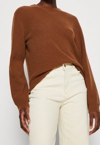 Suéter marrón de punto acanalado con mangas largas, cuello redondo y puños. Combinado con jeans claros ajustados.