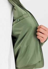 Jachetă bomber de culoare verde măslinie, cu o textură netedă, având o închidere cu fermoar, manșete ribbă și un buzunar cu capse în interior.