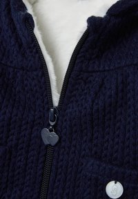 Giacca zip-up blu navy con esterno a maglia testurizzata, morbido rivestimento bianco, cursore a forma di cuore e due tasche frontali.