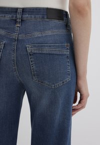 Blue denim jeans with a smooth texture, featuring a leather brand patch at the waistband and two back pockets with topstitching details.