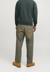 Uomo che indossa jeans blu sbiaditi, stivali in suede beige e una felpa verde scuro, in piedi con la schiena rivolta alla fotocamera su uno sfondo semplice.