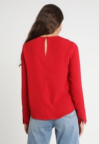 Blusa rossa a maniche lunghe con apertura a chiave sulla schiena, tessuto liscio e polsini con bottoni. Abbinata a jeans chiari a vita alta.