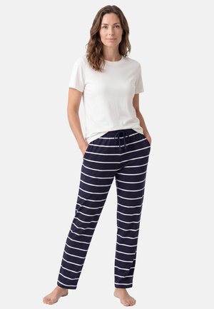 Donna in piedi a piedi nudi, che indossa una camicia bianca a maniche corte e pantaloni blu navy con righe orizzontali bianche, mani in tasca.