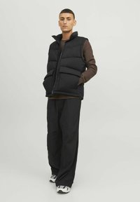 Schwarze gepolsterte Weste mit hohem Kragen, ausgestattet mit zwei Fronttaschen und einem Reißverschluss auf der Vorderseite. Getragen über einem braunen Langarmshirt und schwarzen Hosen.