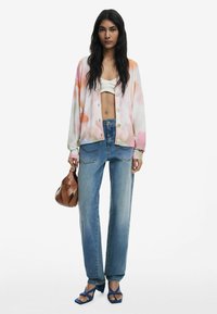 Bunt gestreifter Cardigan mit rosa und orangefarbenen Spritzern, bauchfreies weißes Top, gerade geschnittene blaue Jeans, blaue Riemchensandalen und eine braune Handtasche.