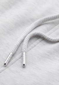 Gros plan de cordons de capuche blancs tressés avec des embouts métalliques gravés de lettres sur un tissu gris clair.