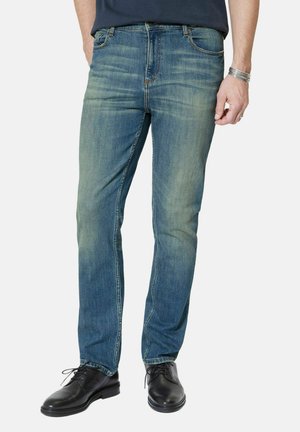 Man draagt vervaagde blauwe spijkerbroek, zwarte veterlaarzen en een marineblauw overhemd, staat met één hand ontspannen langs zijn zijde.