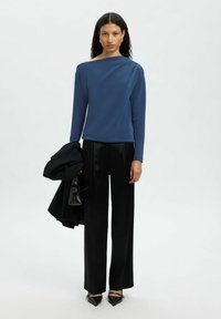 Langärmliges blaues Oberteil mit asymmetrischem Ausschnitt, kombiniert mit hoch taillierten schwarzen Hosen und schwarzen Pumps mit spitzer Zehenform.