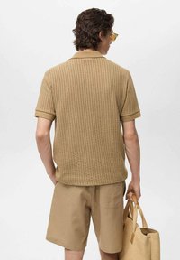 Polo shirt a coste color beige chiaro con maniche corte, abbinato a pantaloni corti beige chiaro. Porta una borsa intrecciata beige, vista da dietro.