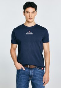 Jonge man met kort donker haar, gekleed in een marineblauw T-shirt met de tekst "Authentic" en een blauwe spijkerbroek, staand met één hand in de zak tegen een effen achtergrond.
