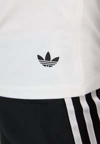 Balta medvilninė marškinėliai su juodu Adidas logotipu, kuriame yra trys lapai, ir dvi horizontaliu juostelės apačioje kairėje pusėje. Juodos kelnės su baltomis juostelėmis.