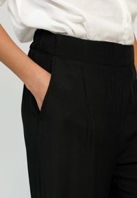 Pantalon noir en tissu léger avec une taille élastique. Doté d'une poche latérale et de plis subtils, associé à une chemise blanche.