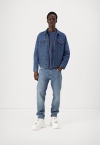 Μπλε τζιν μπουφάν με δύο μπροστινές τσέπες, φορεμένο πάνω από μπλε μπλουζάκι, συνδυασμένο με ανοιχτό μπλε τζιν και λευκά ογκώδη αθλητικά παπούτσια.