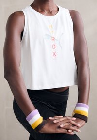 Valkoinen lyhyt tanktoppi, jossa on "HYROX" -grafiikka oranssina, vaaleanpunaisena ja sinisenä. Mustat kuvioidut legginsit ja värikkäät rannekkeet purppuran, keltaisen ja ruskean sävyissä.