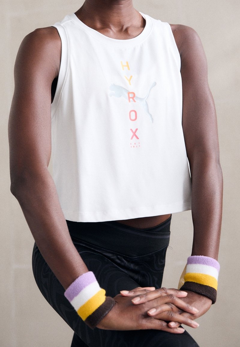 Valkoinen lyhyt tanktoppi, jossa on "HYROX" -grafiikka oranssina, vaaleanpunaisena ja sinisenä. Mustat kuvioidut legginsit ja värikkäät rannekkeet purppuran, keltaisen ja ruskean sävyissä.