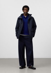 Man som bär en mörk marinblå hooded jacka över en blå pikétröja, vida marinblå byxor och svarta slip-on skor, stående mot en enkel bakgrund.